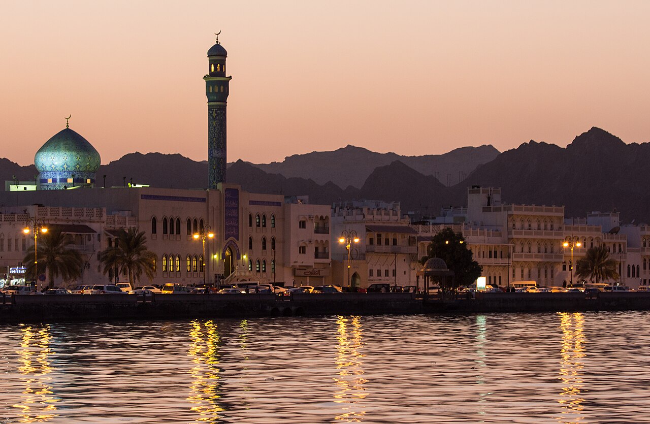 Mutrah Corniche in Muscat at dusk: a lit mosque and minaret against the Hajar mountains, reflected in the harbour